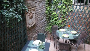 Un patio al aire libre con una mesa preparada para dos, rodeado de sillas de mimbre, un muro de ladrillo y una valla metálica decorativa.