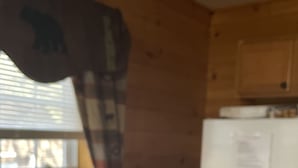 A kitchen with wooden walls, a window with blinds, a refrigerator, and a chair.