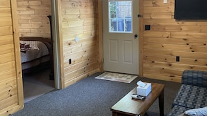 A wooden-paneled room with a door, a bed, a small table with a tissue box, and a chair.