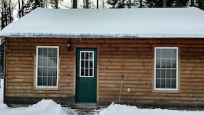 A wooden cabin with a green door, two windows, and a snow-covered roof.