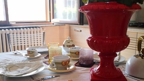 Ein Esstisch mit einer Platte Wurstwaren und Käse, eine rote Vase mit Blumen und ein Fenster mit Jalousien.