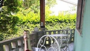 Ein Tisch mit einer Champagnerflasche und einem Glas auf einer gemusterten Tischdecke, platziert auf einer Terrasse mit Metallstühlen und einem Geländer.
