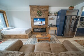 A living room with a fireplace, a TV, a sofa, a bed, and a kitchen area. - Modern Studio with Balcony & Stunning Views! (Keystone)