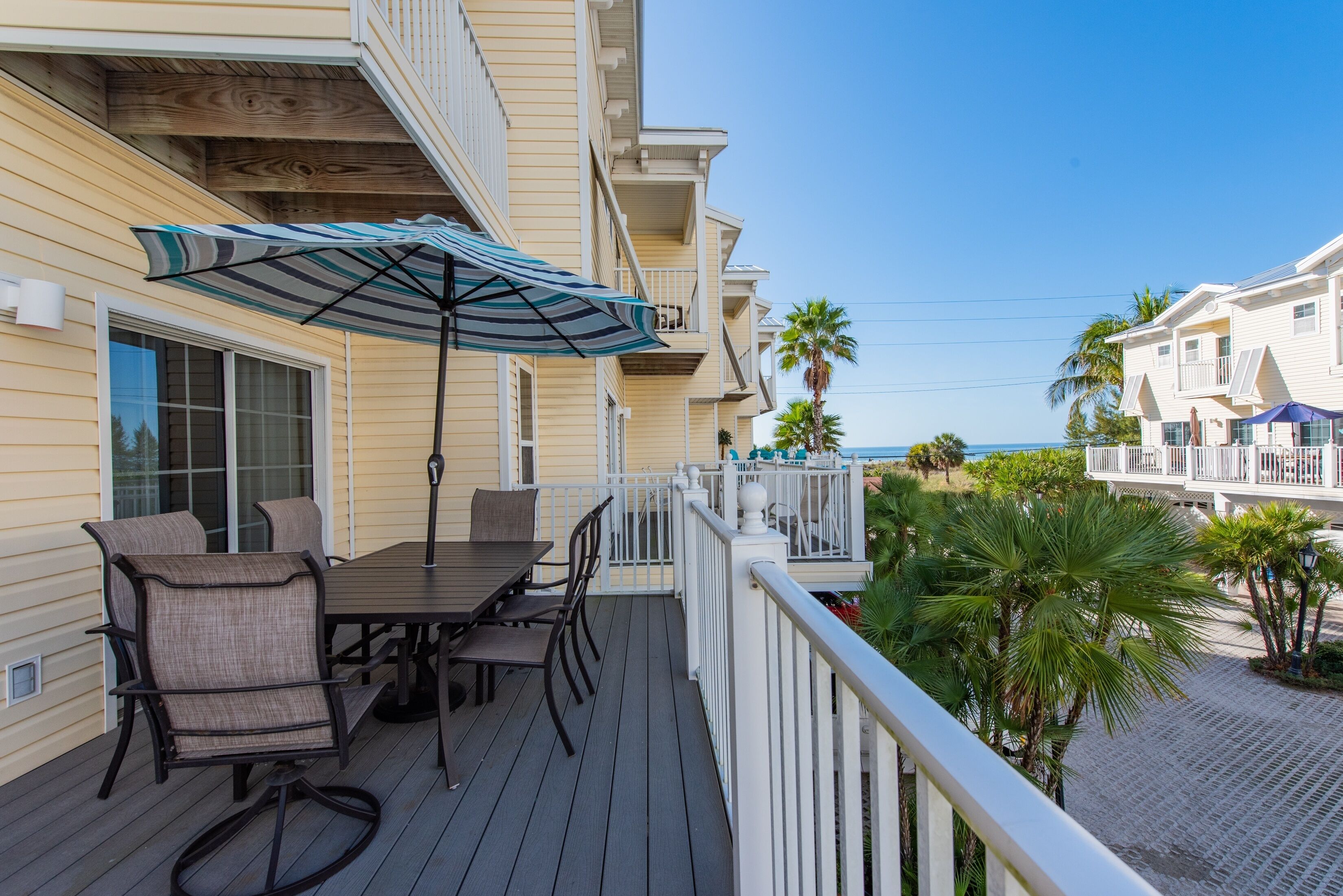 Um deck de madeira com mesa e cadeiras, um guarda-sol listrado e vista para a praia e o oceano.