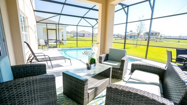 An outdoor patio area with wicker furniture, a glass-top table, and a view of a swimming pool and grassy area.