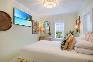 A bedroom with a bed, a television, a window with blinds, and wall-mounted artwork.