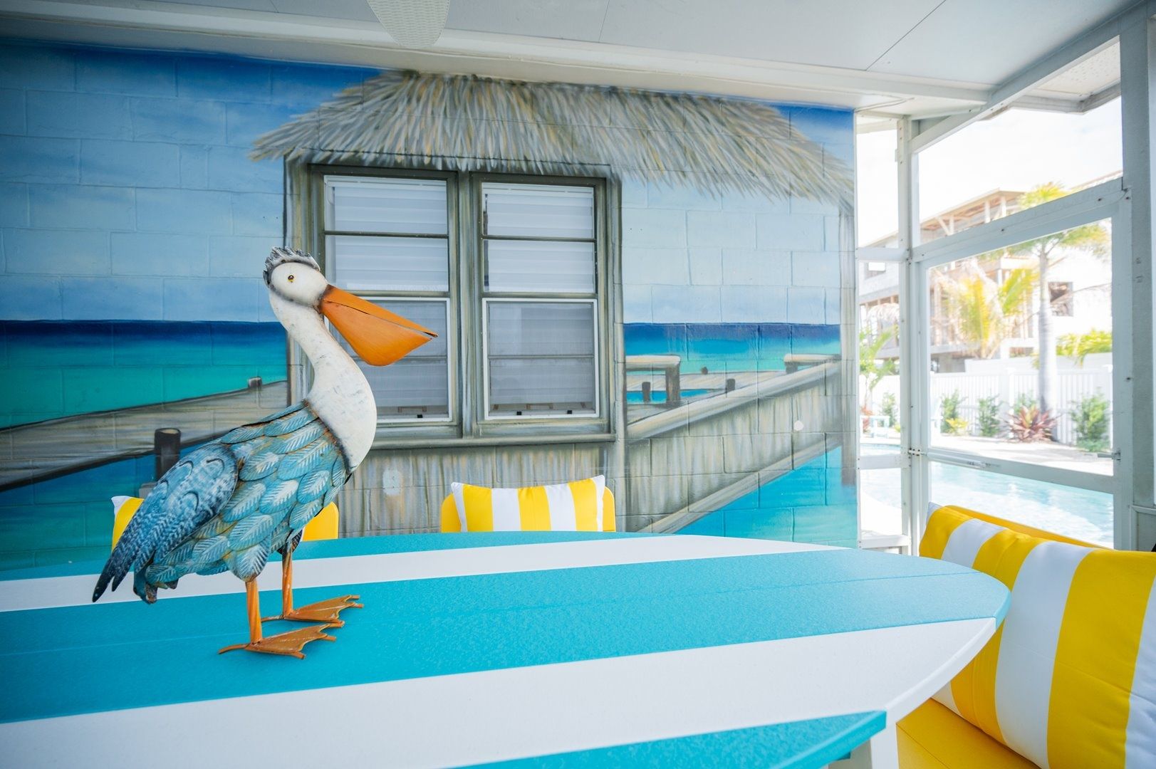 A room with a large window overlooking a pool, a thatched-roof structure, and a table with striped tablecloths.