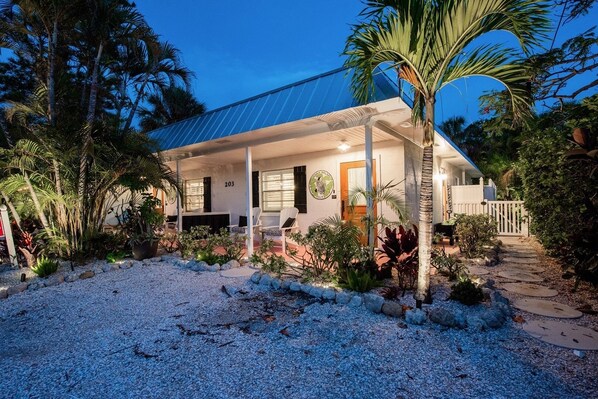 A house with a covered porch, number 203, surrounded by tropical plants and a gravel driveway. - Sandpail 203 71st St West 2 Bedroom Villa by RedAwning (Holmes Beach)