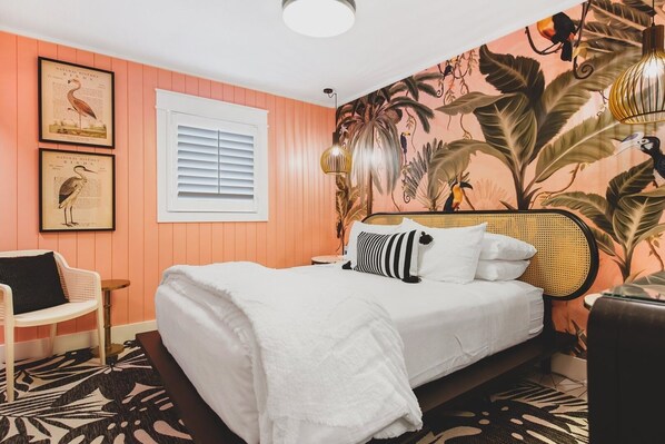 A bedroom with a bed, a chair, a window with blinds, and a wall with a tropical mural. - Sandbox 203 71st St East 2 Bedroom Villa by RedAwning (Holmes Beach)
