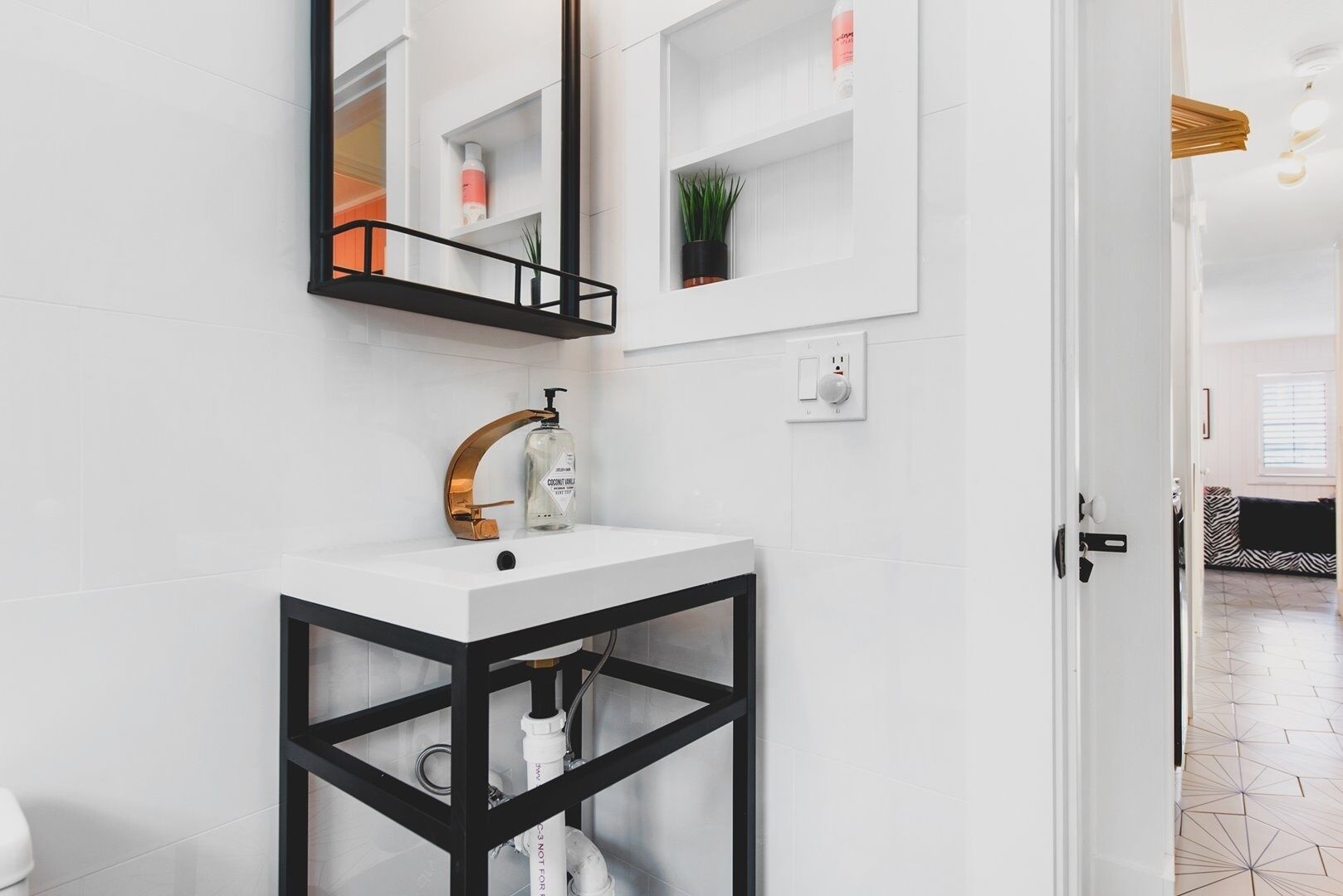 A modern bathroom with a white sink, black frame, and a large mirror. There is a shelf with toiletries and a potted plant.