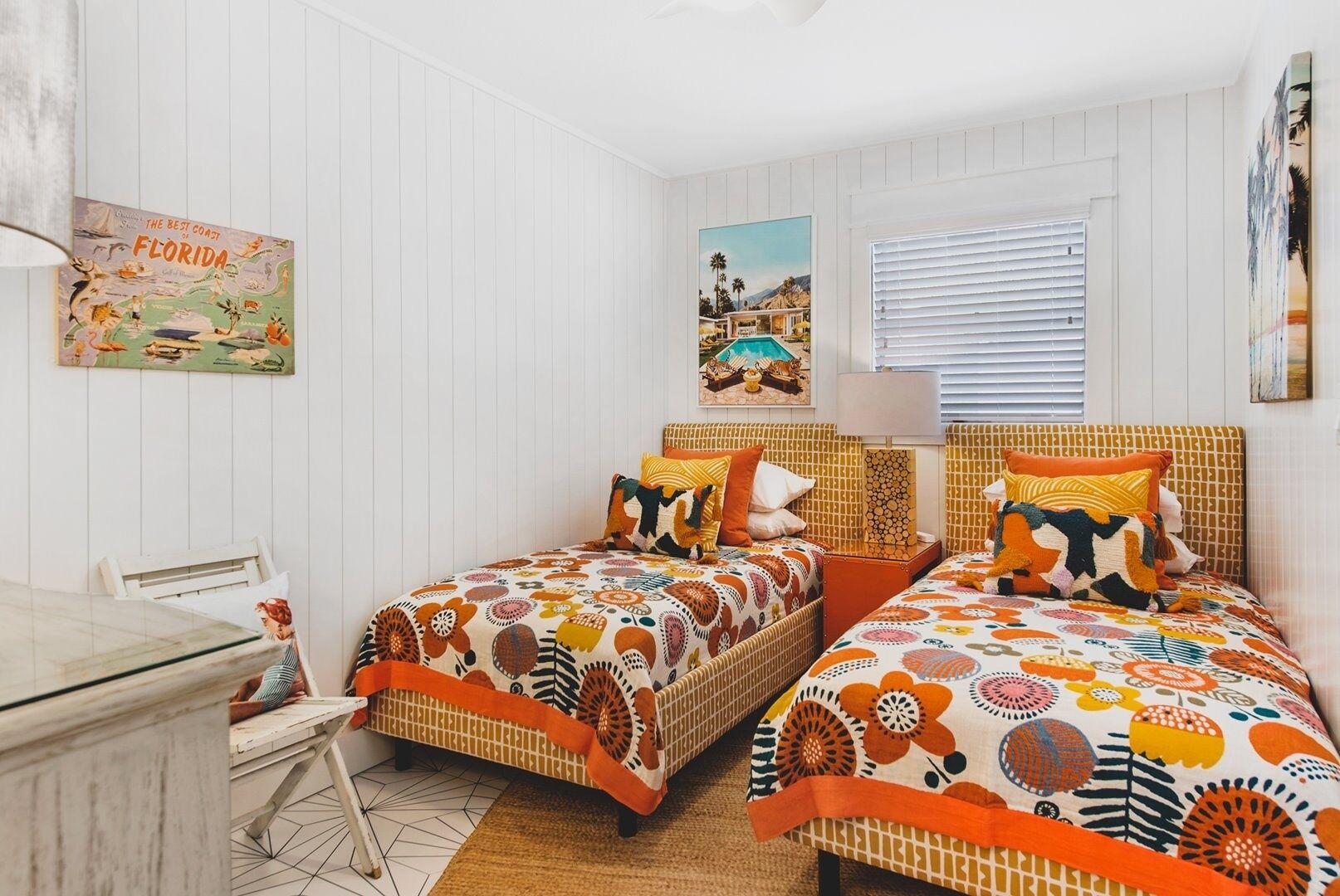 A bedroom with two beds, a desk, and a chair. There are wall-mounted pictures and a window with blinds.