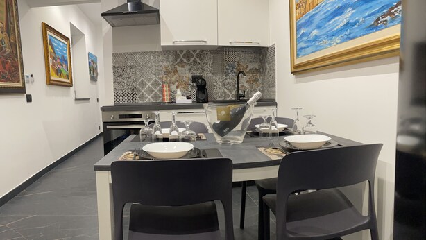 Une cuisine moderne avec une table à manger dressée pour deux personnes, comprenant des chaises noires, un saladier blanc et des verres à vin.