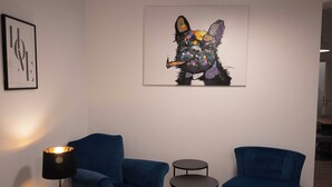 Une pièce avec deux fauteuils bleus, une petite table et une lampe. Une œuvre d’art aux couleurs vives est accrochée au mur.