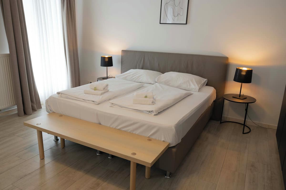 A neatly made bed with white linens, two bedside tables with lamps, a wooden bench, and a framed artwork on the wall.