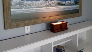 A framed painting of a beach scene on a wall, a small wooden box, and a white shelf below. - Just  a 2 minute walk to the Atlantic Ocean!
 (Surf City)