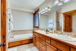 A bathroom with a bathtub, two sinks, wooden cabinets, a large mirror, and a ceiling light fixture. - Snow Chateau - Ski In/Out|Walk to Main St|Hot Tub (Breckenridge)