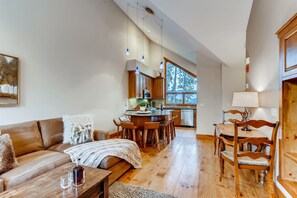 A spacious living area with a sofa, coffee table, and dining table with chairs. A kitchen is visible in the background with a large window. - Snow Chateau - Ski In/Out|Walk to Main St|Hot Tub (Breckenridge)