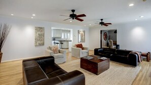 A living room with a brown leather sofa, a white armchair, a black leather sofa, and a wooden coffee table. There are two ceiling fans, a kitchen in the background, and a painting on the wall.