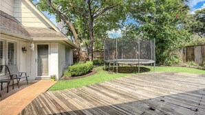 A wooden deck with a trampoline, surrounded by a fenced yard with trees and a house.