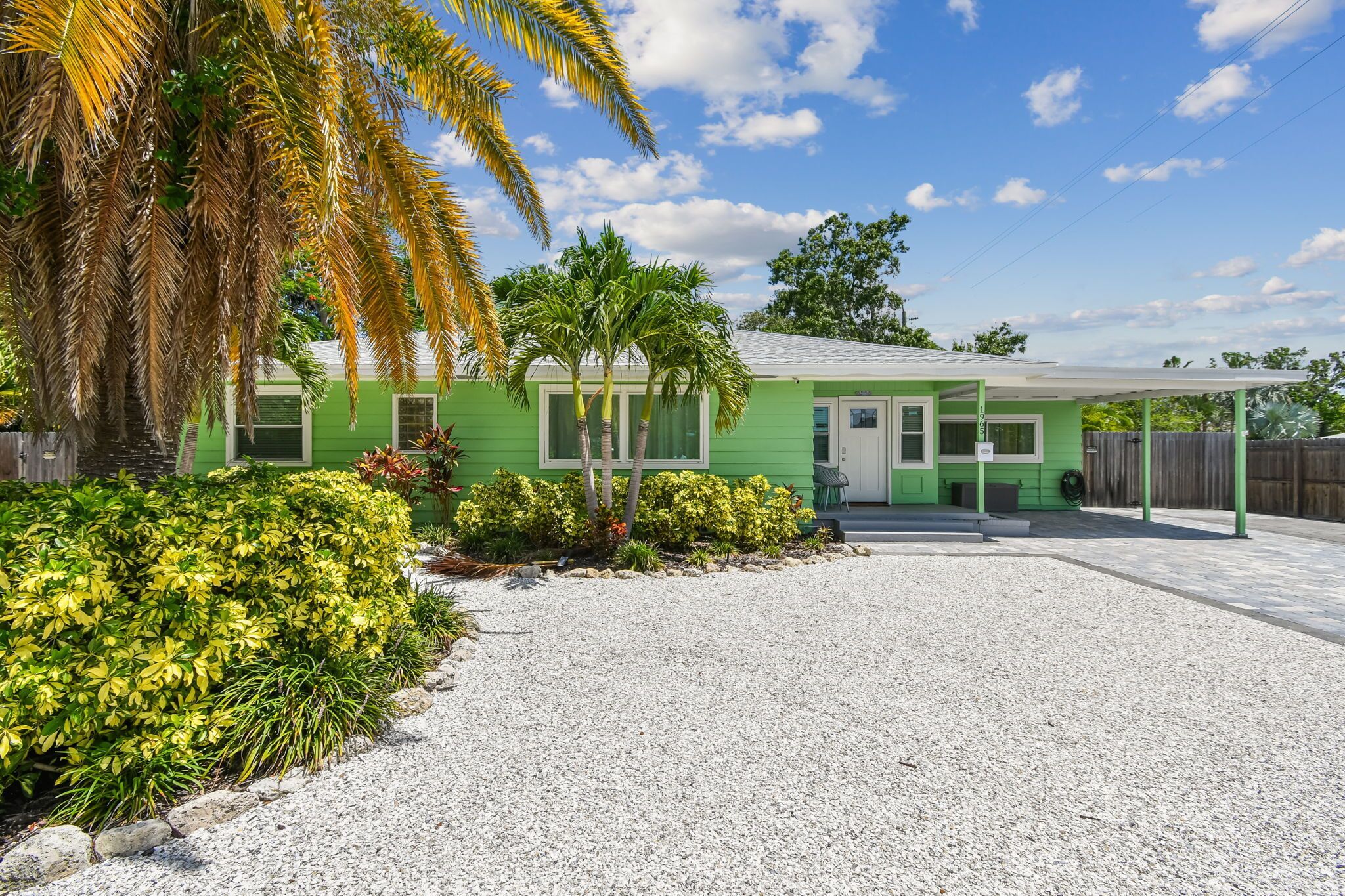 A green house with a gravel driveway, palm trees, and a fenced yard.