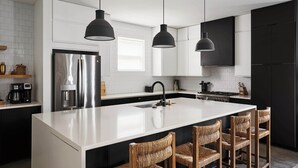 A modern kitchen with a central island, black pendant lights, stainless steel appliances, and wicker bar stools. - Stylish 3-Bed Retreat, Rooftop, Parking, Sleeps 10  |  Ensuite Baths, Bunk Room, Gourmet Kitchen, Cozy Living Space, Rooftop Views & Fire Table, Near Downtown, Distilleries & Marathon Village, 24 Hr Hospitality (Nashville)