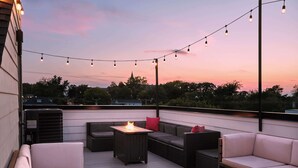 An outdoor seating area with a fire table, cushioned sofas, and string lights against a sunset backdrop. - Stylish 3-Bed Retreat, Rooftop, Parking, Sleeps 10  |  Ensuite Baths, Bunk Room, Gourmet Kitchen, Cozy Living Space, Rooftop Views & Fire Table, Near Downtown, Distilleries & Marathon Village, 24 Hr Hospitality (Nashville)