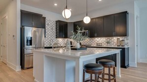 A modern kitchen with dark cabinets, a central island with stools, and a patterned backsplash. - Roomy 1-Level Condo Near Vanderbilt With Parking    |    1 Story, Secure Parking, Patio. 3 miles to Broadway (Nashville)