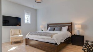 A bedroom with a wooden bed, a chair, a bedside table with a lamp, and a TV mounted on the wall. - Roomy 1-Level Condo Near Vanderbilt With Parking    |    1 Story, Secure Parking, Patio. 3 miles to Broadway (Nashville)