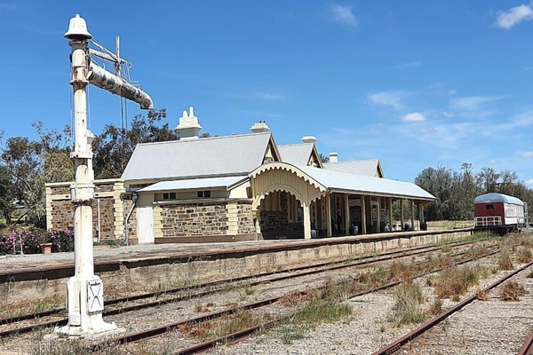 A historic stone railway station with ornate roofing and a wide platform stands beside old, overgrown train tracks under a bright blue sky, with a single red and white train car in the distance.