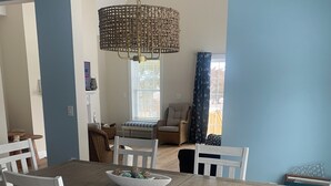 A dining area with a wooden table and white chairs, a wicker chandelier, and a view of the outdoors through a window with curtains. - New construction. Short Walk to Beautiful Ridegevale Beach (Chatham)