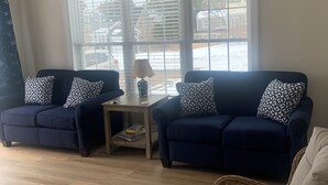 A living room with two dark blue sofas, each with patterned pillows, a small wooden side table, a white pouf, and a wicker chair. - New construction. Short Walk to Beautiful Ridegevale Beach (Chatham)