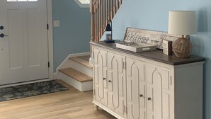A hallway with a wooden staircase, a white door with a window, and a cabinet with a welcome sign. - New construction. Short Walk to Beautiful Ridegevale Beach (Chatham)