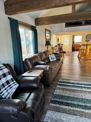 A spacious living room with a leather sofa, a dining area with wooden chairs, and a kitchen visible through an open door. - Cozy cottage across from the Lake (Bay Port)