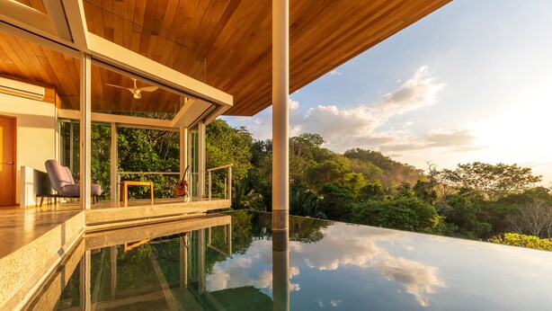 Una casa moderna con un porche de cristal que da a una piscina, rodeada de exuberante vegetación.