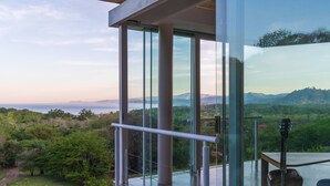 Un balcón moderno de cristal con vistas a un paisaje montañoso y una guitarra sobre una mesa.