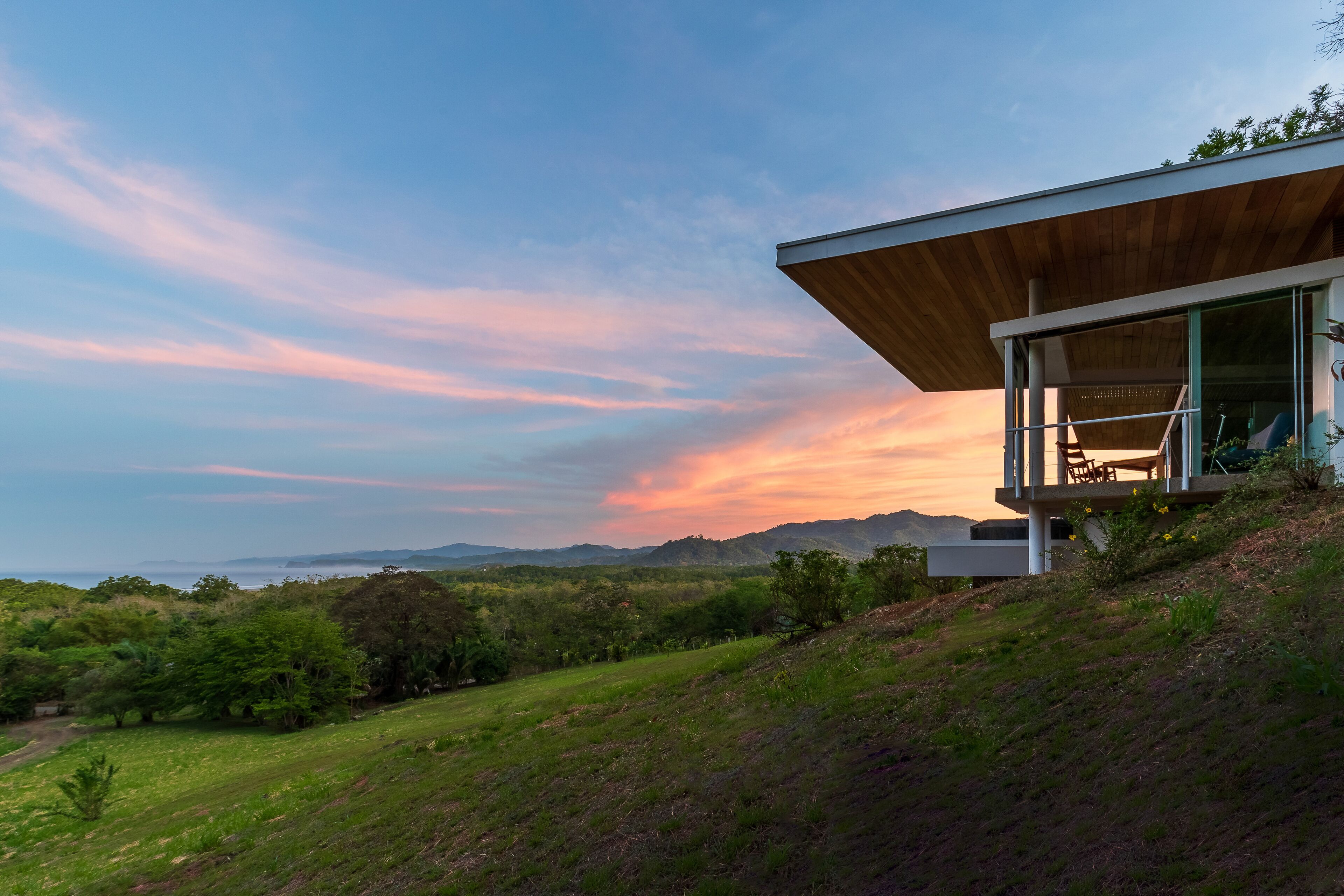 A modern house with a wooden roof and large glass windows, surrounded by lush greenery and hills.
