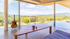 Una habitación moderna con ventanas amplias, techo de madera, una guitarra, una mesa y vistas a un paisaje montañoso.
