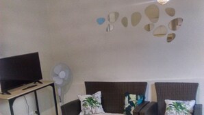 A living room with a flat-screen TV on a stand, a glass coffee table with a green decorative item, two brown sofas with white cushions, and a wall-mounted light fixture. - Dépendance Avec Entrée Privée et Terrasse (Fort-de-France)