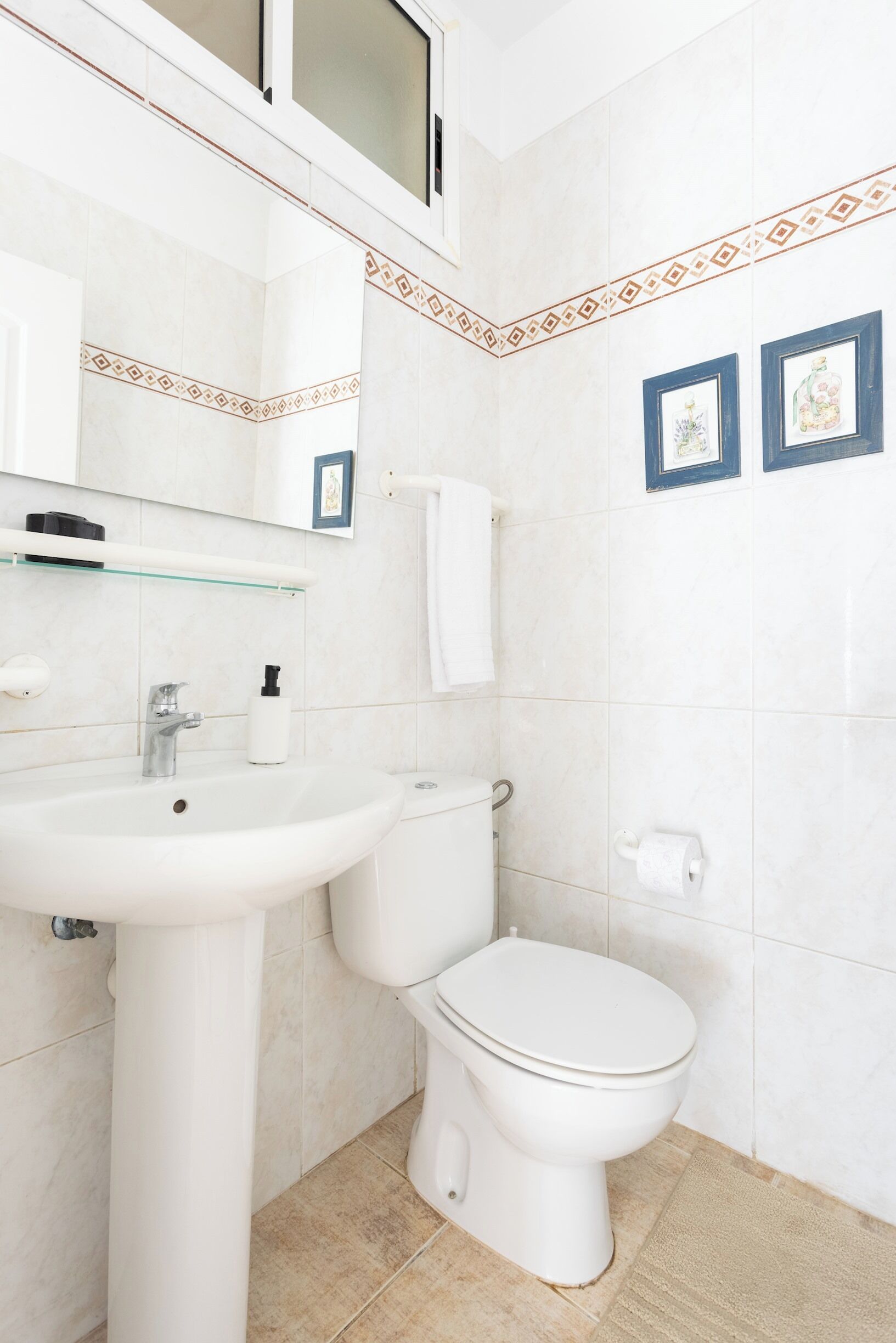 A bathroom with a white sink, toilet, and mirror. There are framed pictures on the wall and a towel hanging on the wall.