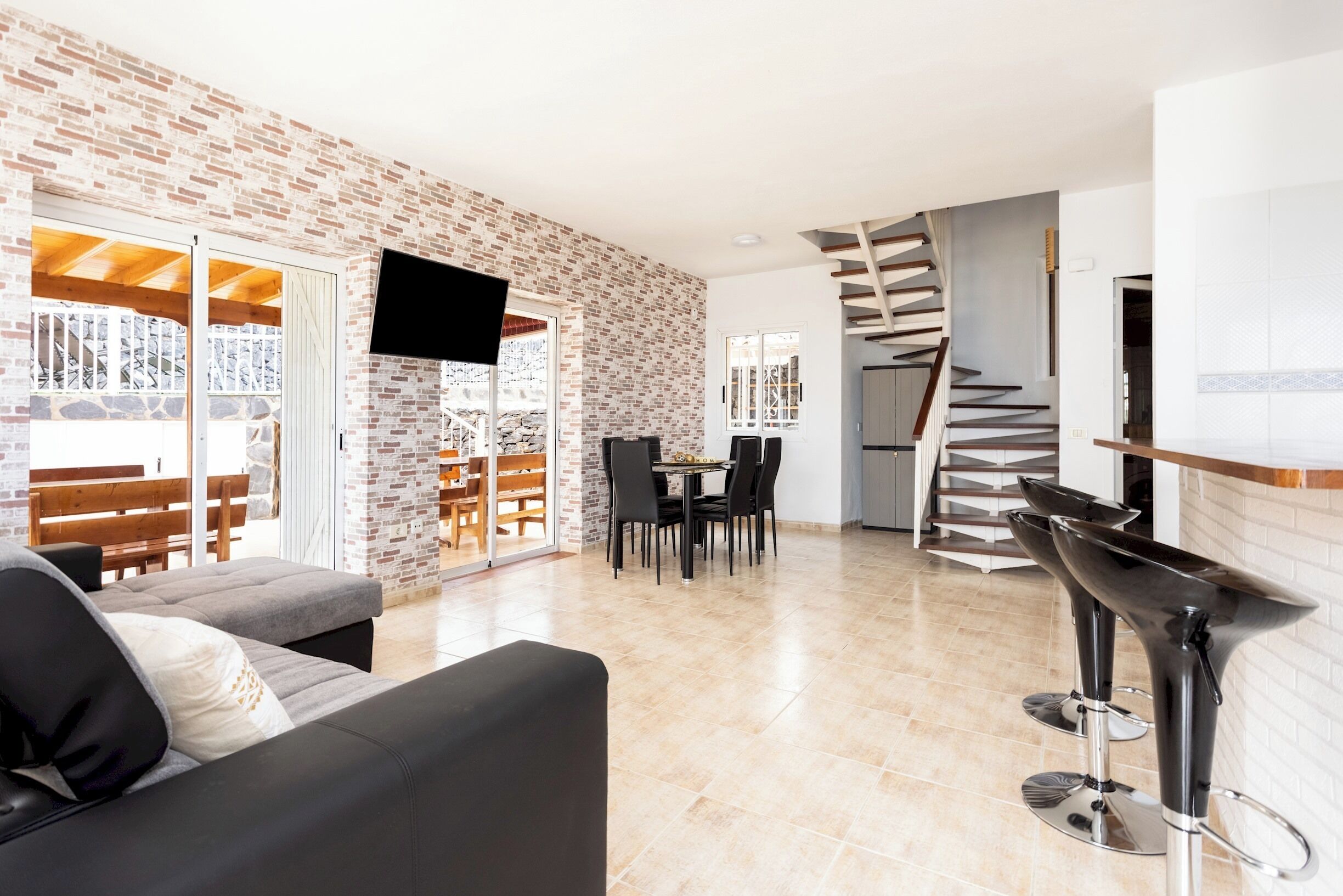 A modern living space with a brick wall, a flat-screen TV, a dining area, and a spiral staircase.