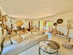 Ein geräumiges Wohnzimmer mit einem großen, beige gefärbten Sofa, einem Glastisch, Korbmöbeln und Blick durch eine Schiebetür auf einen Außenbereich.