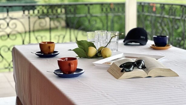 Une table dressée avec des tasses, des verres, un livre et un chapeau, avec en arrière-plan un paysage verdoyant.