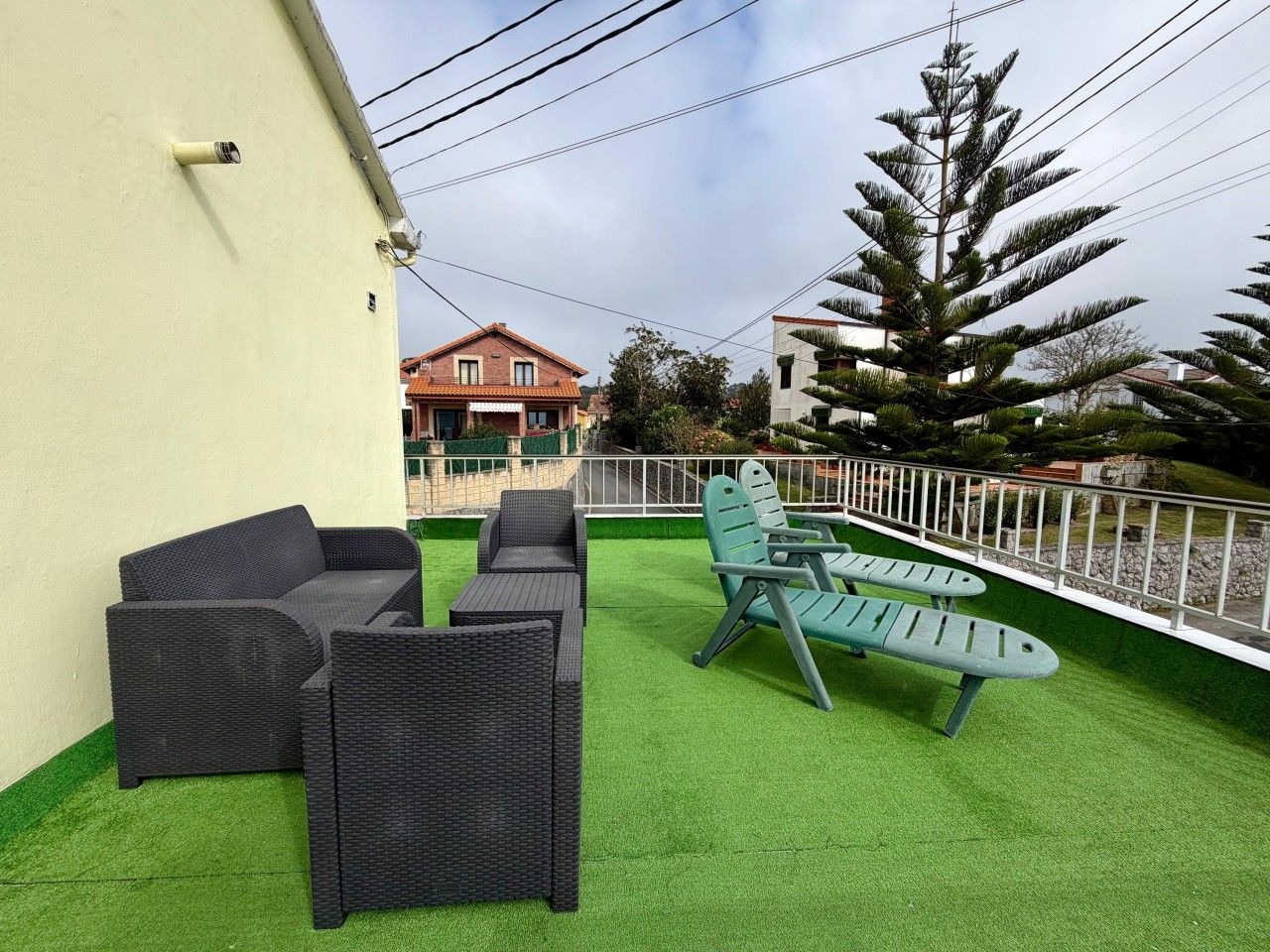 Un área de estar exterior con mobiliario de mimbre y césped artificial, rodeada por una cerca blanca y un árbol grande.