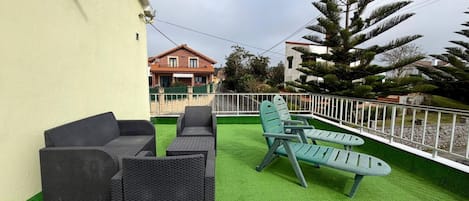 Un área de estar exterior con mobiliario de mimbre y césped artificial, rodeada por una cerca blanca y un árbol grande.