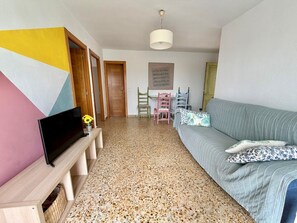Une pièce avec un téléviseur posé sur un meuble en bois, un canapé agrémenté de coussins à motifs, un mur orné d’un motif géométrique et un luminaire au plafond.