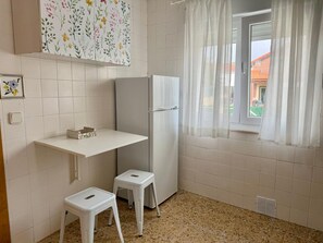 Une cuisine compacte avec des carreaux blancs, un plan de travail blanc, un réfrigérateur, un petit tabouret et une fenêtre avec des rideaux.