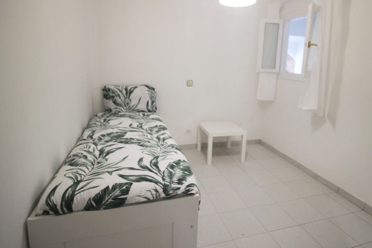 Ein kleines, sauberes Zimmer mit einem Bett, einem kleinen weißen Tisch und einem Fenster mit Vorhängen.