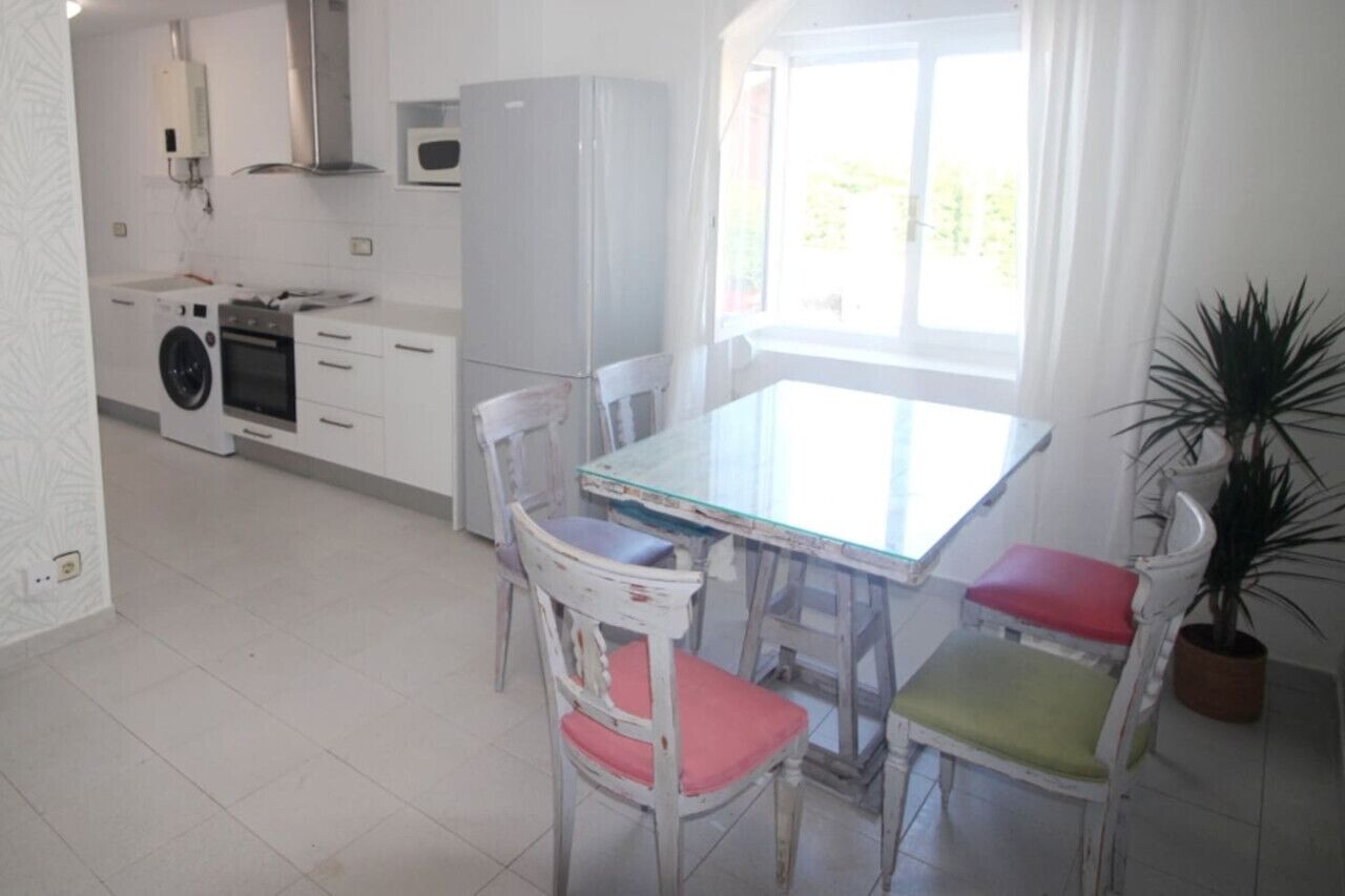 Une cuisine compacte équipée d’un lave-linge, d’un four et d’un four à micro-ondes. On y trouve une table à manger avec quatre chaises, dont une avec un coussin rouge. Une plante en pot est visible dans un coin.