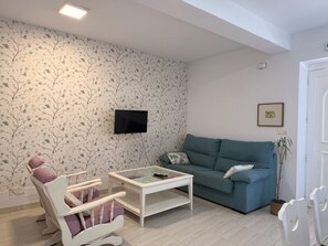 A living room with a teal sofa, a white armchair with cushions, a glass-top coffee table, a wall-mounted TV, and a potted plant.
