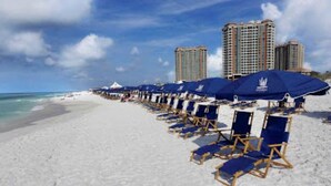 Ein Strand mit blauen Liegestühlen und Sonnenschirmen, ein sandiger Uferstreifen und hohe Gebäude im Hintergrund.